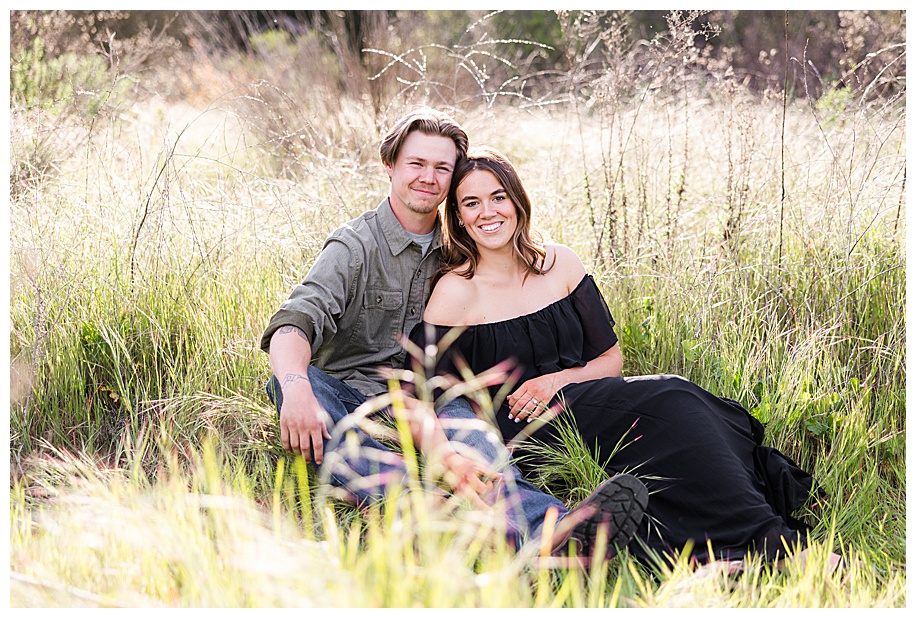 engaged couple sitting in tall grass laughing at the camera